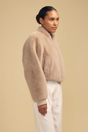 A person wearing a cropped beige ARIANNA FAUX FUR BOMBER JACKET by Velvet by Graham & Spencer with ribbed cuffs and white trousers.