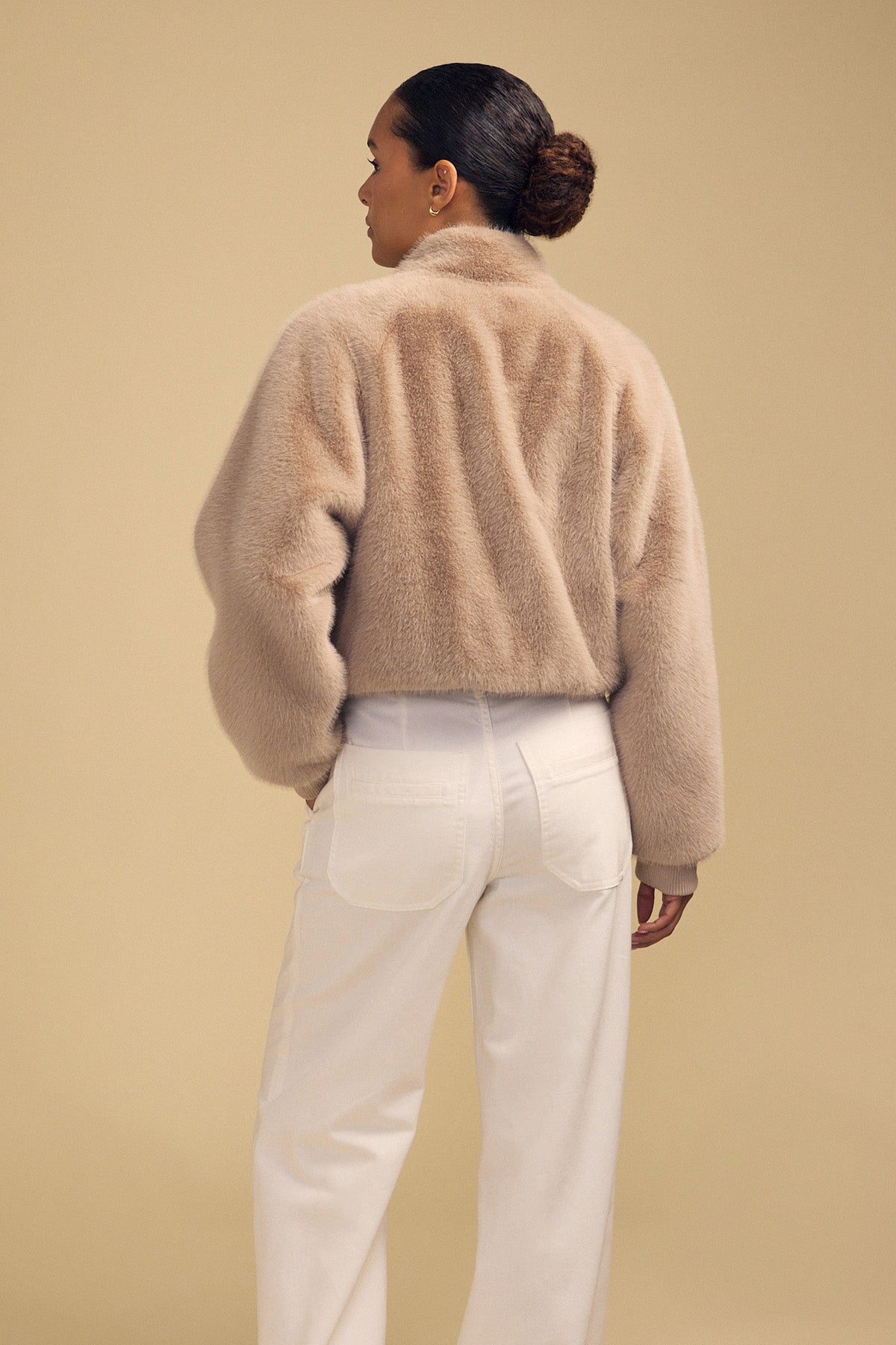 A person seen from behind, wearing an ARIANNA FAUX FUR BOMBER JACKET in beige and white trousers.-46488090214593
