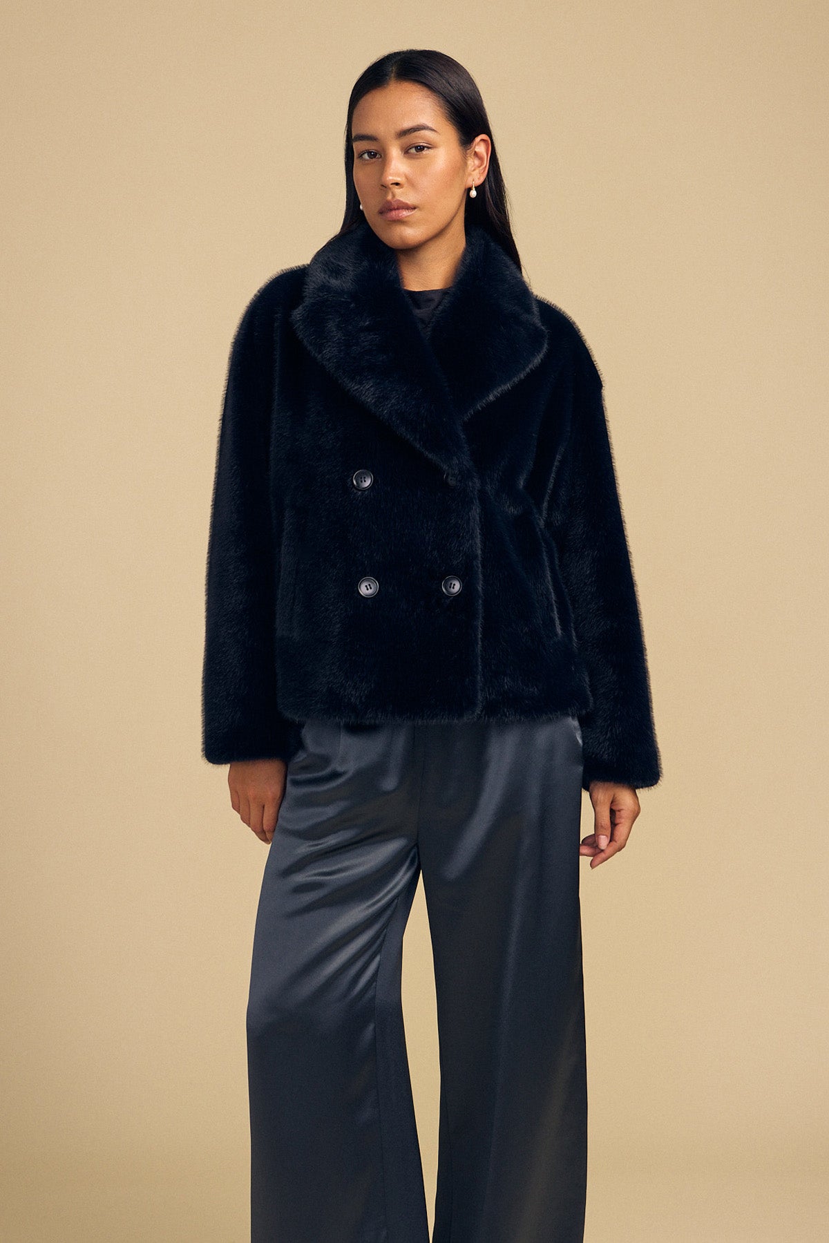 A person wearing a black, double-breasted CHARLOTTE FAUX FUR JACKET as a statement layer paired with dark satin trousers.