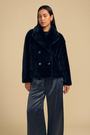 A person wearing a black, double-breasted CHARLOTTE FAUX FUR JACKET as a statement layer paired with dark satin trousers.