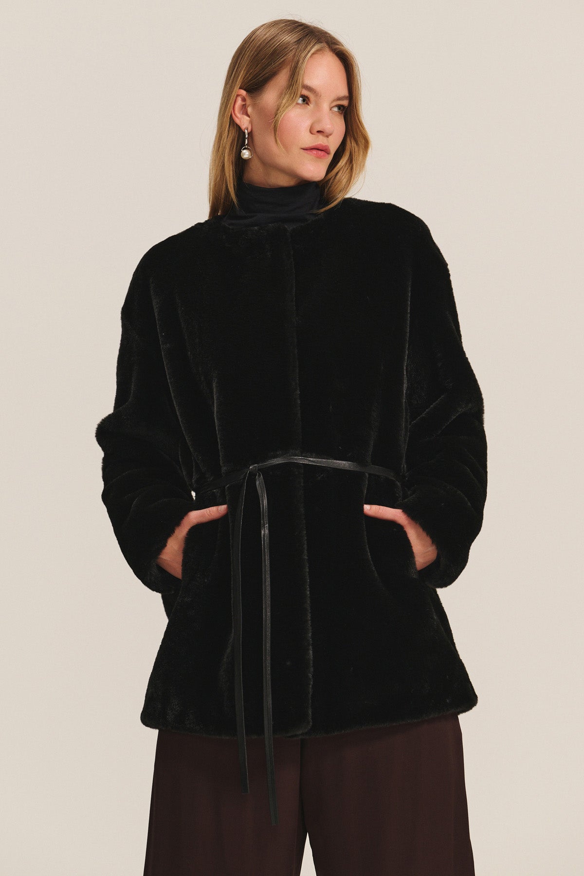 A woman in the MARMONT LUXE FAUX FUR JACKET by Velvet by Graham & Spencer, featuring a detachable belt, dark pants, and drop earrings stands against a light background.-40248194597057