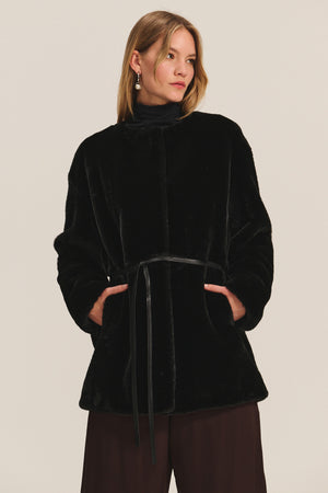 A woman in the MARMONT LUXE FAUX FUR JACKET by Velvet by Graham & Spencer, featuring a detachable belt, dark pants, and drop earrings stands against a light background.
