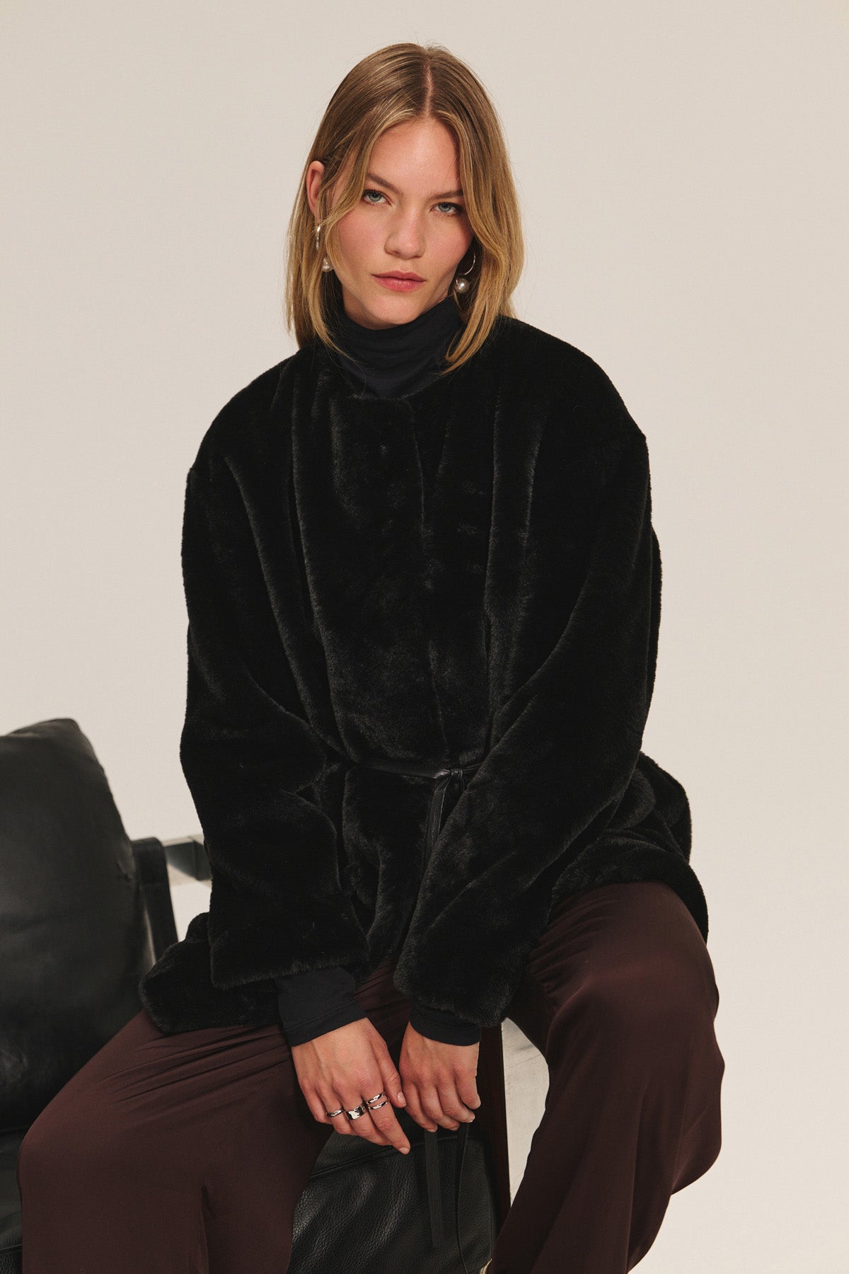 A person with shoulder-length hair wears the Velvet by Graham & Spencer MARMONT LUXE FAUX FUR JACKET, featuring snap button closures, over a black turtleneck and dark brown pants, seated on a black chair against a plain background.-40248194564289
