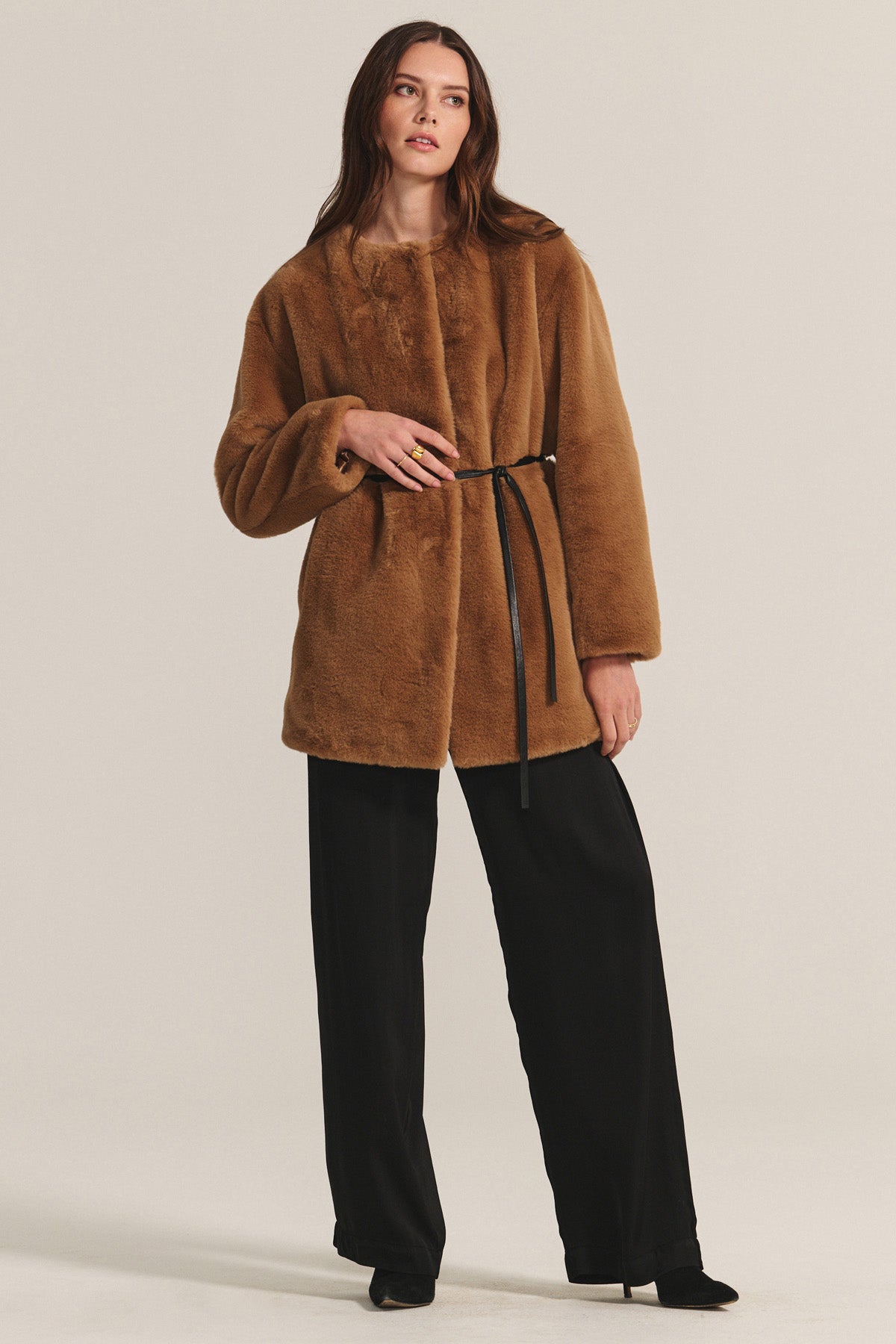   A woman models the MARMONT LUXE FAUX FUR JACKET by Velvet by Graham & Spencer, featuring snap button closures and a thin black belt, styled with black wide-leg trousers and shoes against a plain backdrop. 