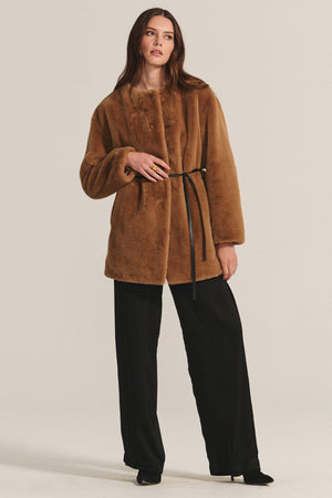 A woman models the MARMONT LUXE FAUX FUR JACKET by Velvet by Graham & Spencer, featuring snap button closures and a thin black belt, styled with black wide-leg trousers and shoes against a plain backdrop.