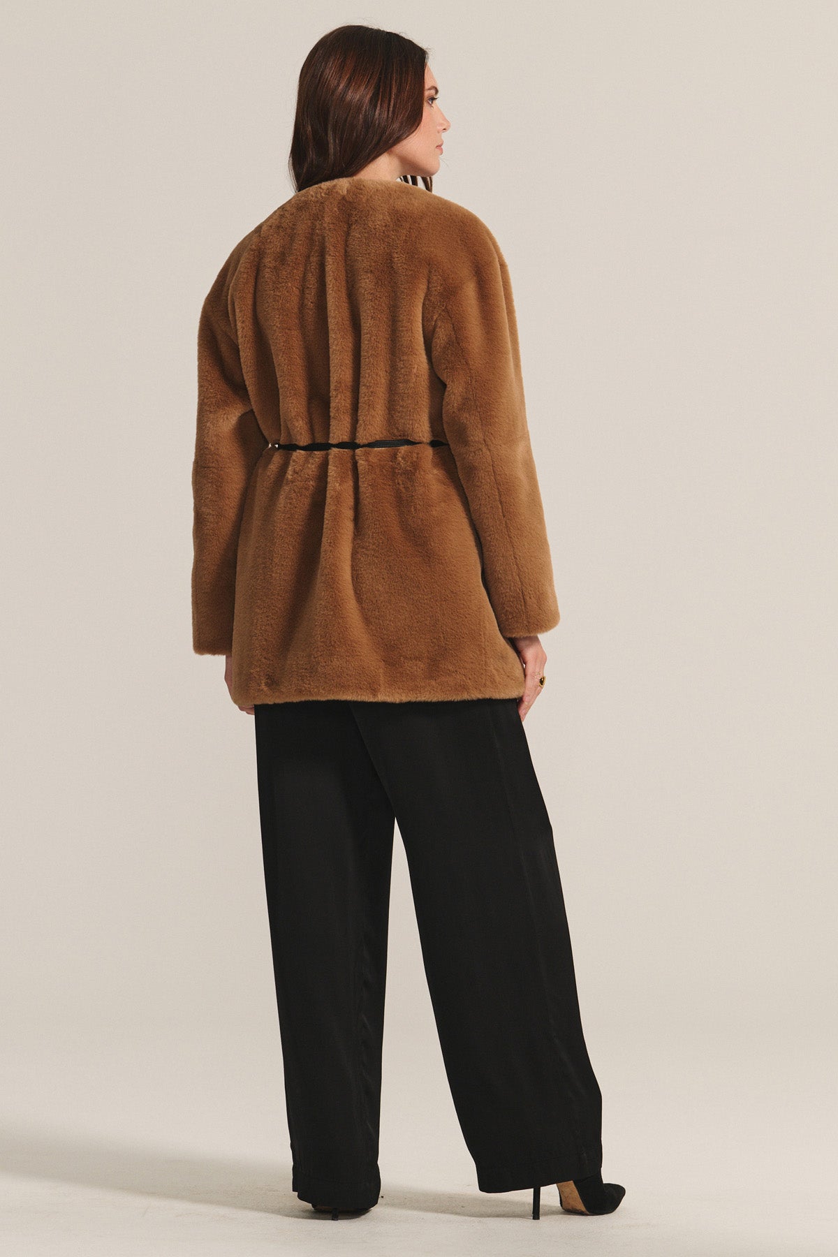 Someone faces away, wearing the MARMONT LUXE FAUX FUR JACKET by Velvet by Graham & Spencer—a light brown faux fur piece with snap buttons and a detachable belt—paired with loose black trousers and black heels.-40248194695361