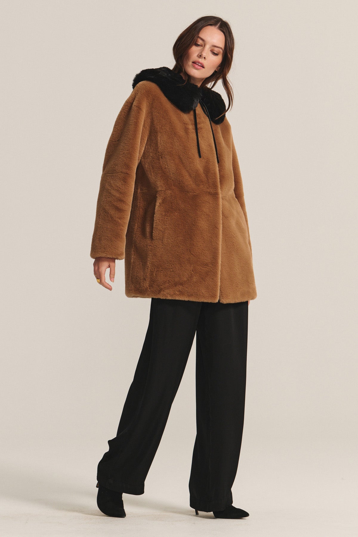 A woman wears the MARMONT LUXE FAUX FUR JACKET by Velvet by Graham & Spencer, featuring snap buttons and a black fur-trimmed hood, paired with black wide-leg trousers and heeled boots against a plain light background.-40248194760897