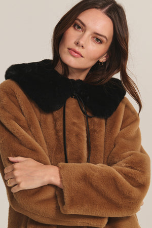 Model wears the MARMONT LUXE FAUX FUR JACKET by Velvet by Graham & Spencer, featuring brown faux fur, a black faux fur collar, and snap button closures, with her arms crossed while looking at the camera.