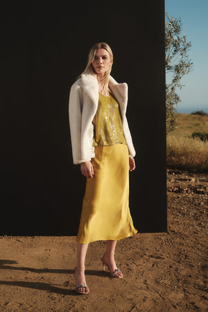 A woman in a yellow skirt and white jacket showcases her chic Behati Sequin Tank Top by Velvet by Graham & Spencer, featuring a scoop neckline and sequin detailing.