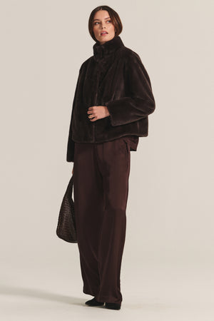 A woman wears the SHEENA LUXE FAUX FUR JACKET by Velvet by Graham & Spencer, paired with wide-leg brown trousers, black shoes, and carries a textured handbag against a plain background.