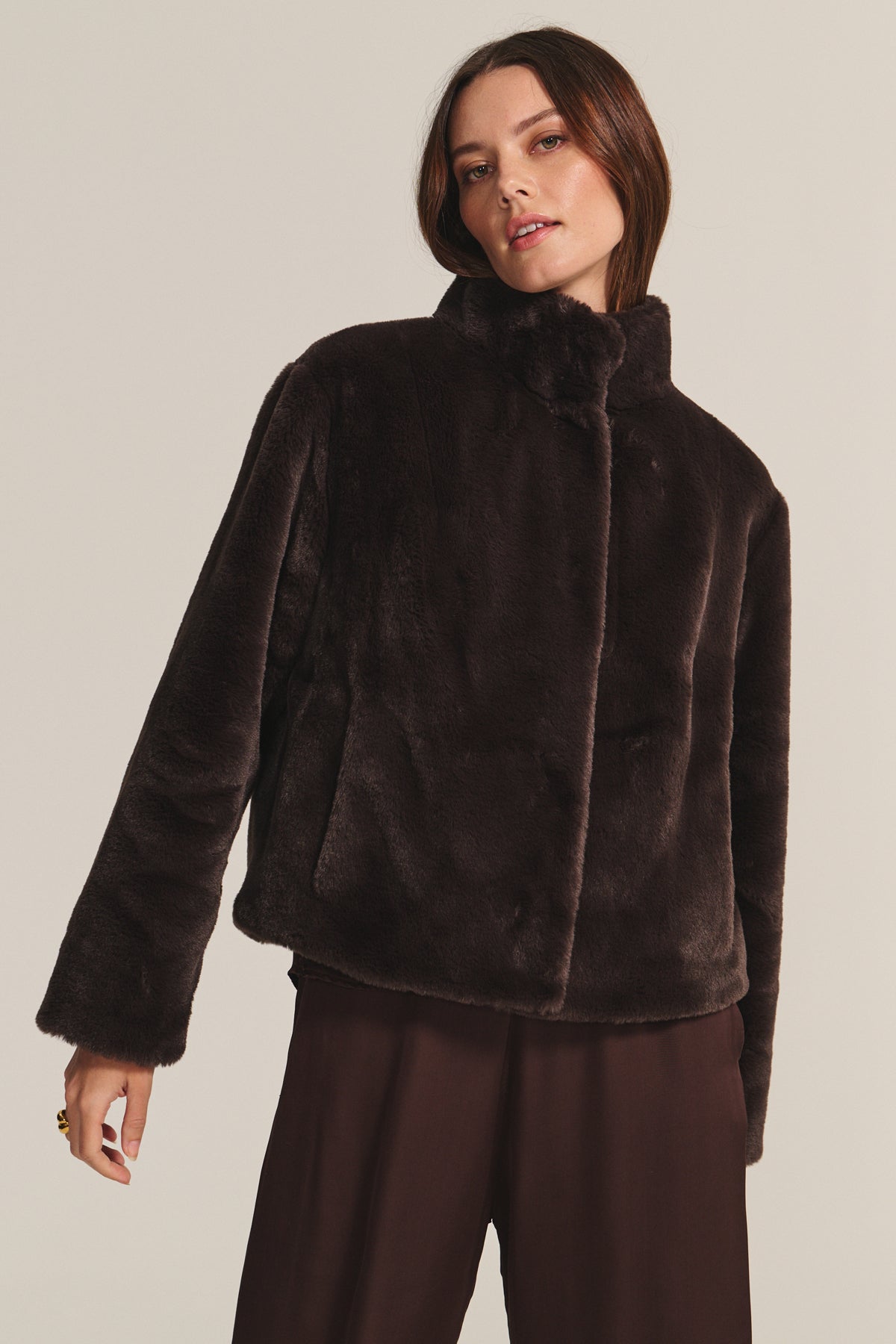 A woman models the SHEENA LUXE FAUX FUR JACKET by Velvet by Graham & Spencer in dark brown with matching pants, set against a plain beige background.-40234934960321