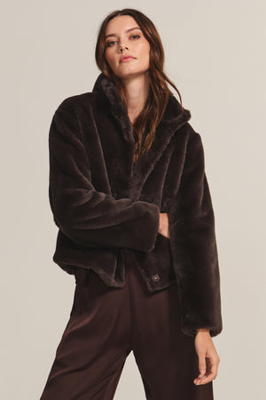 A woman wears the SHEENA LUXE FAUX FUR JACKET by Velvet by Graham & Spencer, a dark brown cozy-fit piece, paired with brown pants against a plain beige background.
