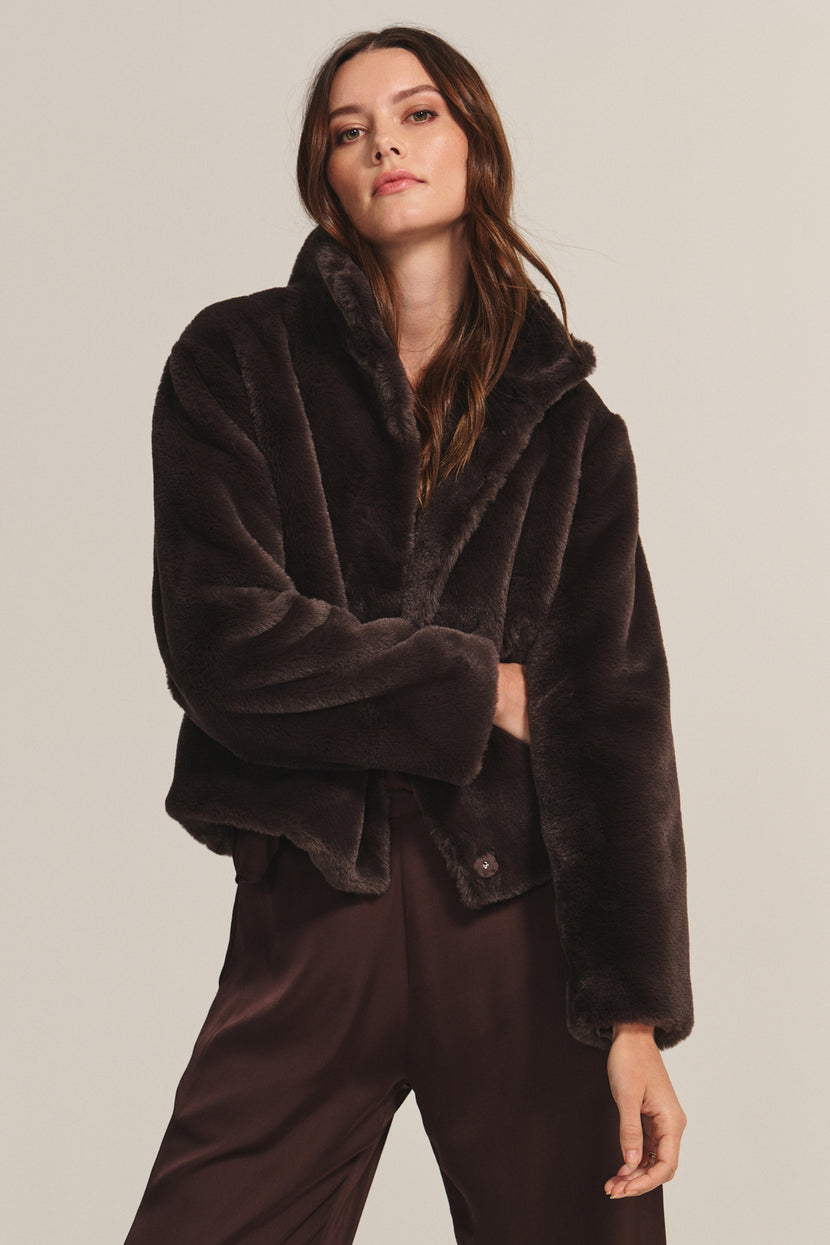 A woman wears the SHEENA LUXE FAUX FUR JACKET by Velvet by Graham & Spencer, a dark brown cozy-fit piece, paired with brown pants against a plain beige background.