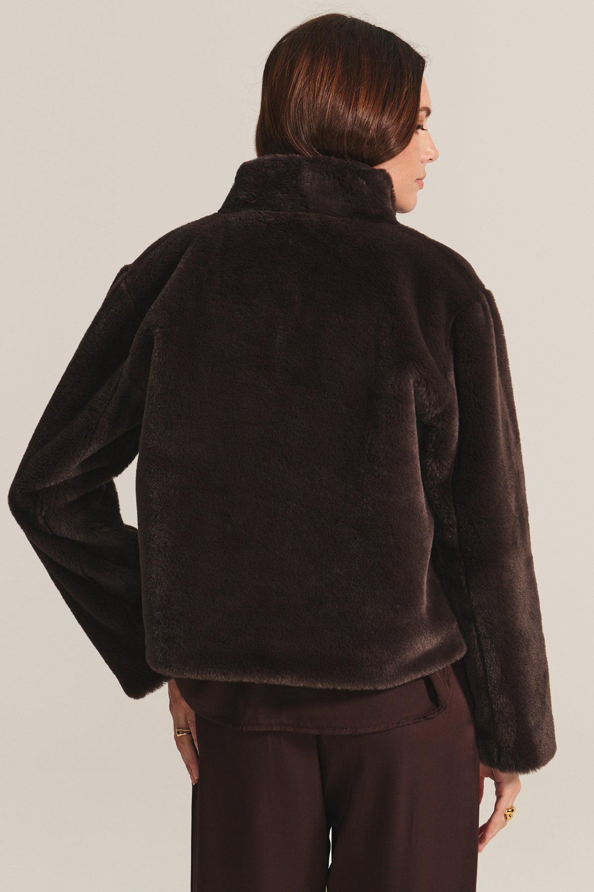 A woman with brown hair models the SHEENA LUXE FAUX FUR JACKET by Velvet by Graham & Spencer and brown pants, seen from the back against a plain background.-40234934894785