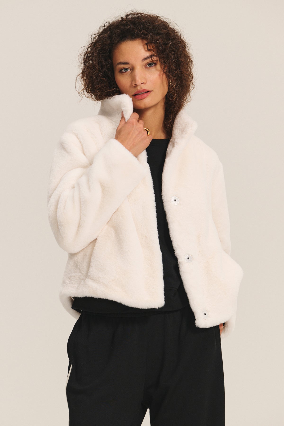   A woman wears the SHEENA LUXE FAUX FUR JACKET by Velvet by Graham & Spencer over a black top and pants, standing against a plain light background. 