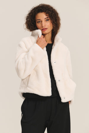 A woman wears the SHEENA LUXE FAUX FUR JACKET by Velvet by Graham & Spencer over a black top and pants, standing against a plain light background.