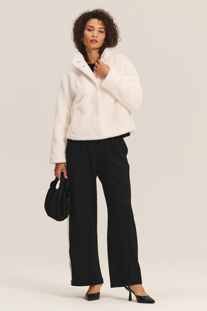 A woman models the SHEENA LUXE FAUX FUR JACKET by Velvet by Graham & Spencer, paired with black wide-leg pants, heels, and a black handbag against a plain background for a chic, cozy look.
