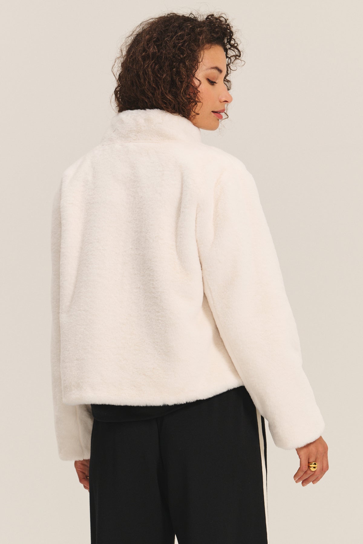 A person with curly hair models the SHEENA LUXE FAUX FUR JACKET by Velvet by Graham & Spencer in white, paired with black pants, showing off its cozy fit while standing against a plain light background facing away from the camera.-40234934796481