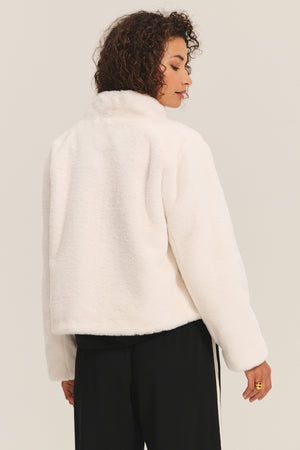 A person with curly hair models the SHEENA LUXE FAUX FUR JACKET by Velvet by Graham & Spencer in white, paired with black pants, showing off its cozy fit while standing against a plain light background facing away from the camera.