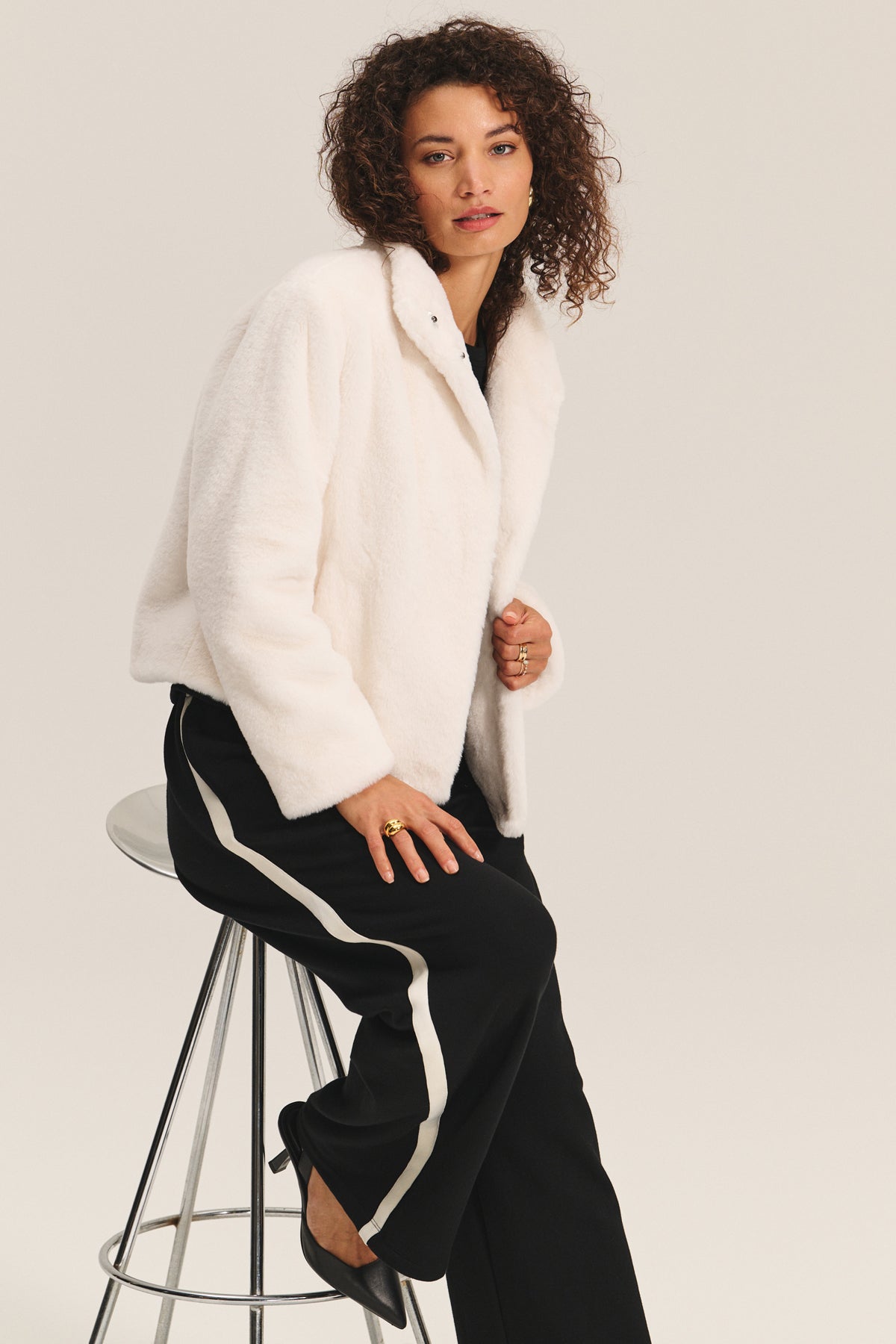   A person with curly hair wears the SHEENA LUXE FAUX FUR JACKET by Velvet by Graham & Spencer, paired with black pants with a white stripe and black shoes, seated on a metal stool against a plain background. 