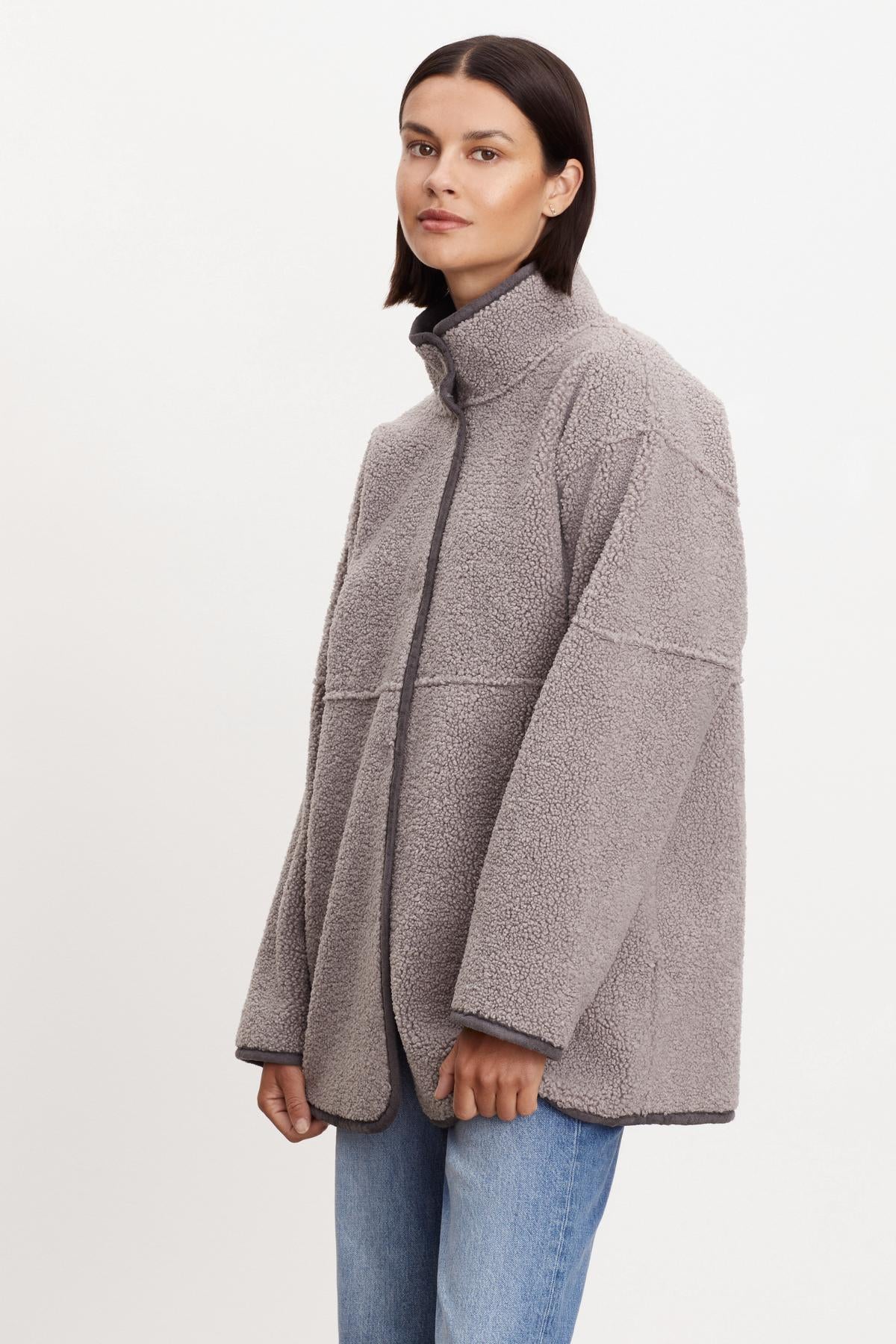 A person wearing a gray ALBANY LUX SHERPA REVERSIBLE JACKET by Velvet by Graham & Spencer and blue jeans stands against a plain white background. The reversible jacket is zipped up and has a high collar.-36094291017921