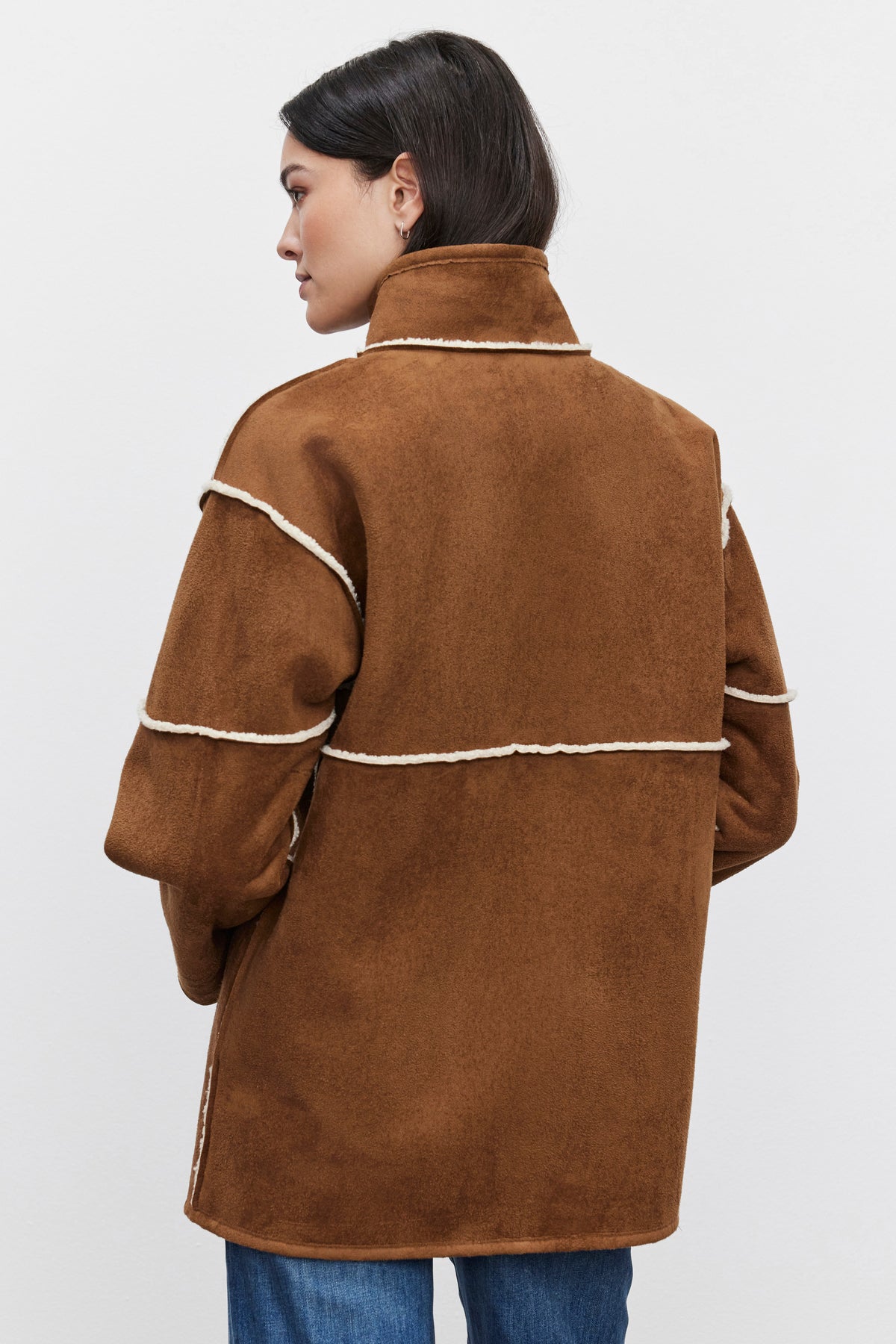   A person with brown hair stands against a plain background, wearing the eco-friendly Albany Luxe Sherpa Reversible Jacket by Velvet by Graham & Spencer, featuring white trim and paired with blue jeans, their back to the camera. 