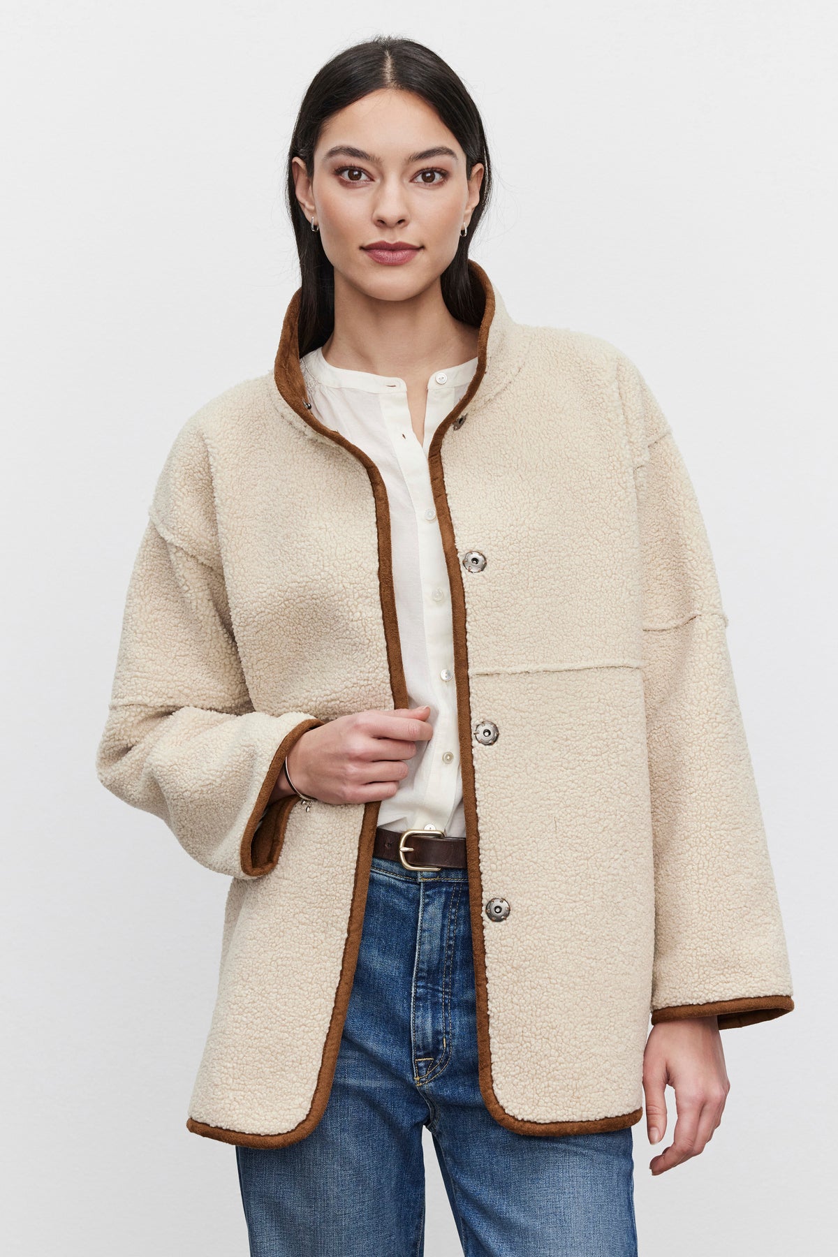   A person clad in the ALBANY LUXE SHERPA REVERSIBLE JACKET by Velvet by Graham & Spencer, featuring a cream-colored, eco-friendly faux sherpa with brown trim, stands against a plain backdrop over a white shirt and blue jeans. 