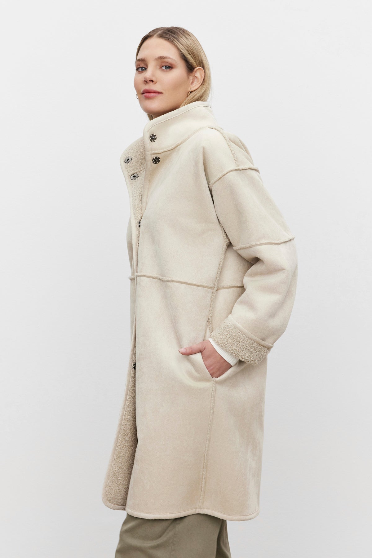   A person wearing a Velvet by Graham & Spencer CARA LUXE SHERPA REVERSIBLE JACKET, showcasing a beige faux suede design with a high collar and one hand in the pocket, stands elegantly against a plain white background. 