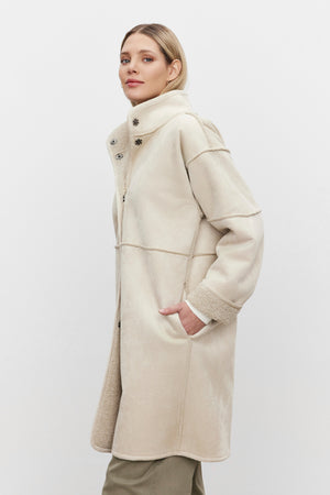 A person wearing a Velvet by Graham & Spencer CARA LUXE SHERPA REVERSIBLE JACKET, showcasing a beige faux suede design with a high collar and one hand in the pocket, stands elegantly against a plain white background.