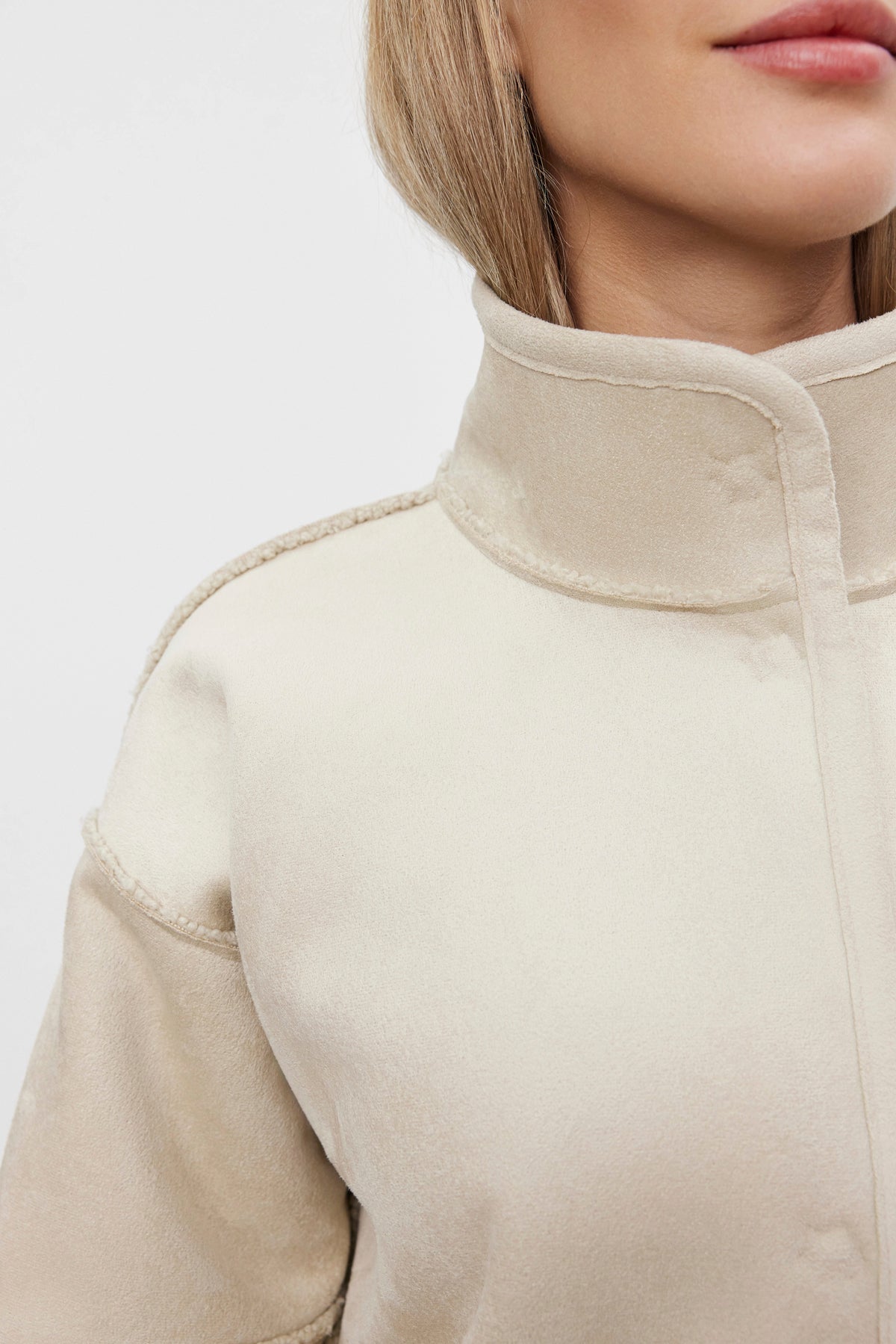   Close-up of a person wearing the CARA LUXE SHERPA REVERSIBLE JACKET in light beige, featuring a high collar and prominent seams. The jacket by Velvet by Graham & Spencer, made with faux suede material, boasts a soft texture against the plain white background. 