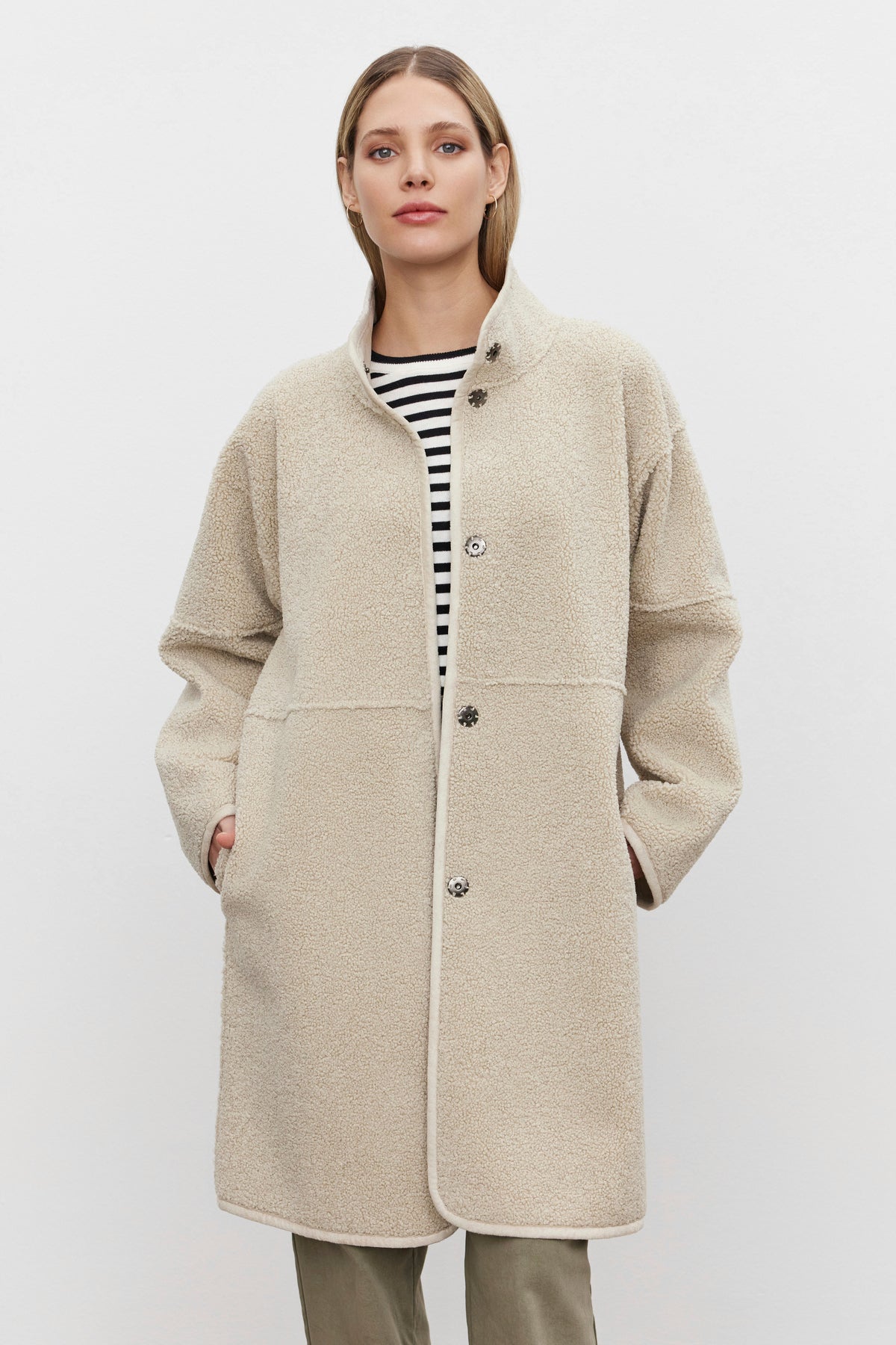   A person wearing a beige wool coat over a black and white striped top stands against a plain background, exuding timeless elegance reminiscent of the Velvet by Graham & Spencer's Cara Luxe Sherpa Reversible Jacket's classic silhouette. 