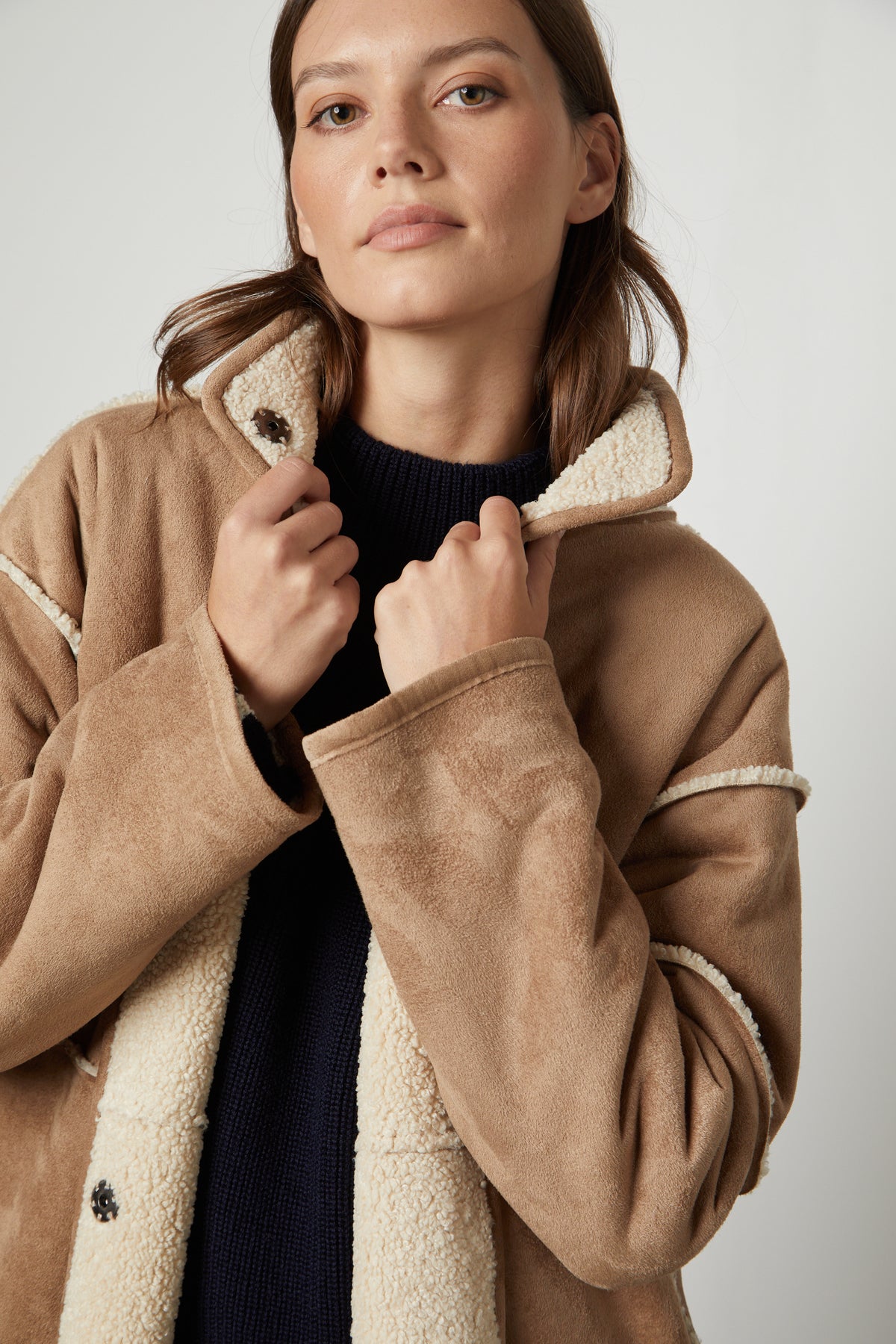 A person wearing the CARA LUXE SHERPA REVERSIBLE JACKET by Velvet by Graham & Spencer, with a black sweater underneath, looks directly at the camera.-26897723982017