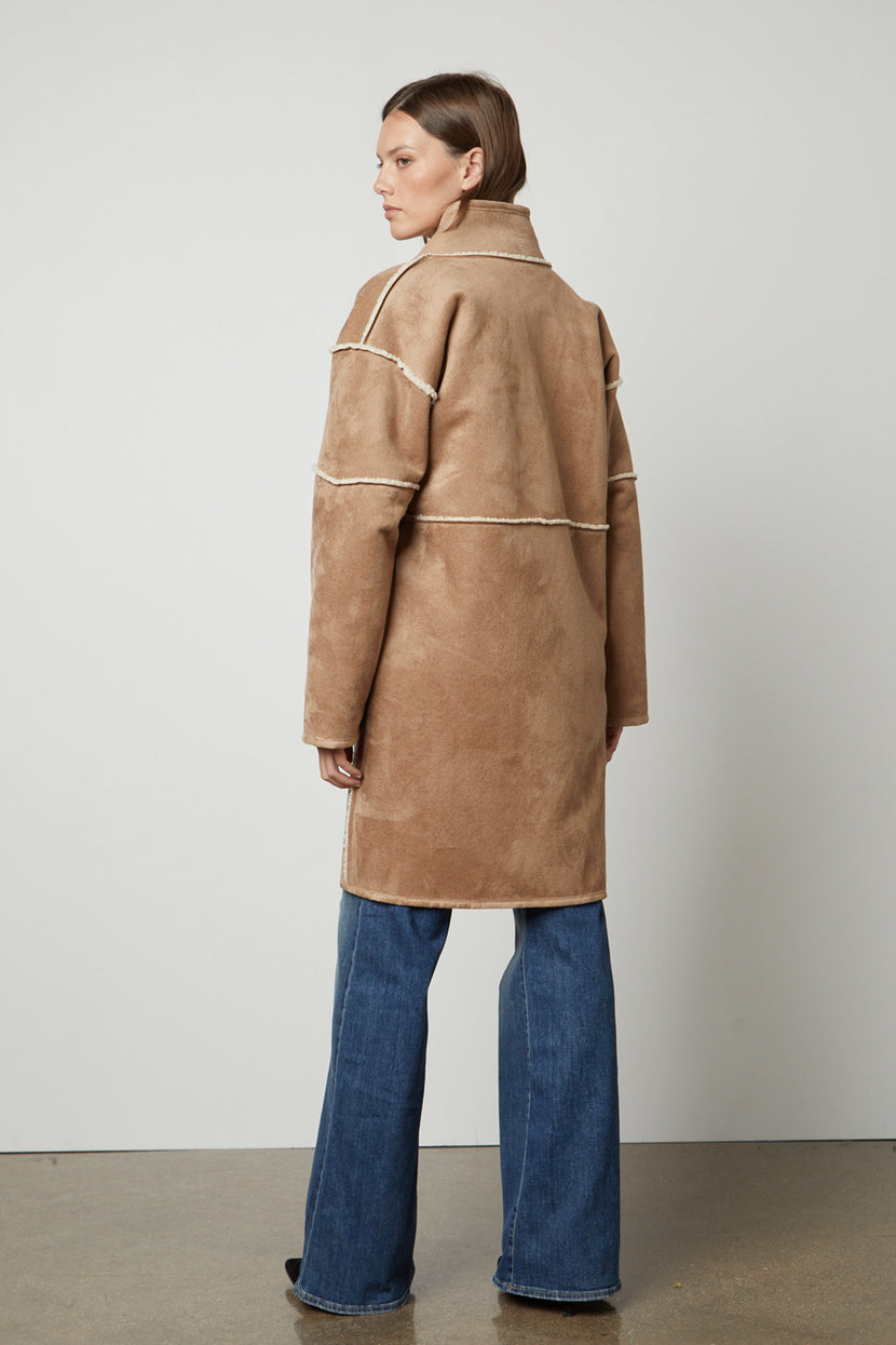 A person wearing a long tan CARA LUXE SHERPA REVERSIBLE JACKET by Velvet by Graham & Spencer and blue jeans stands facing away against a plain background.