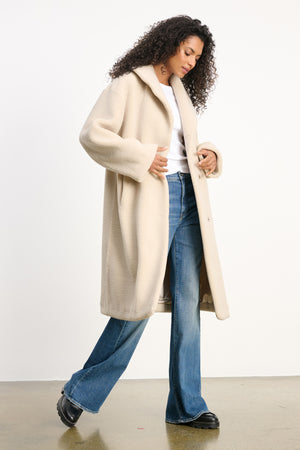 A person wears the ELEANOR SHERPA REVERSIBLE COAT by Velvet by Graham & Spencer over a white top, blue flared jeans, and black shoes, standing against a plain white background.