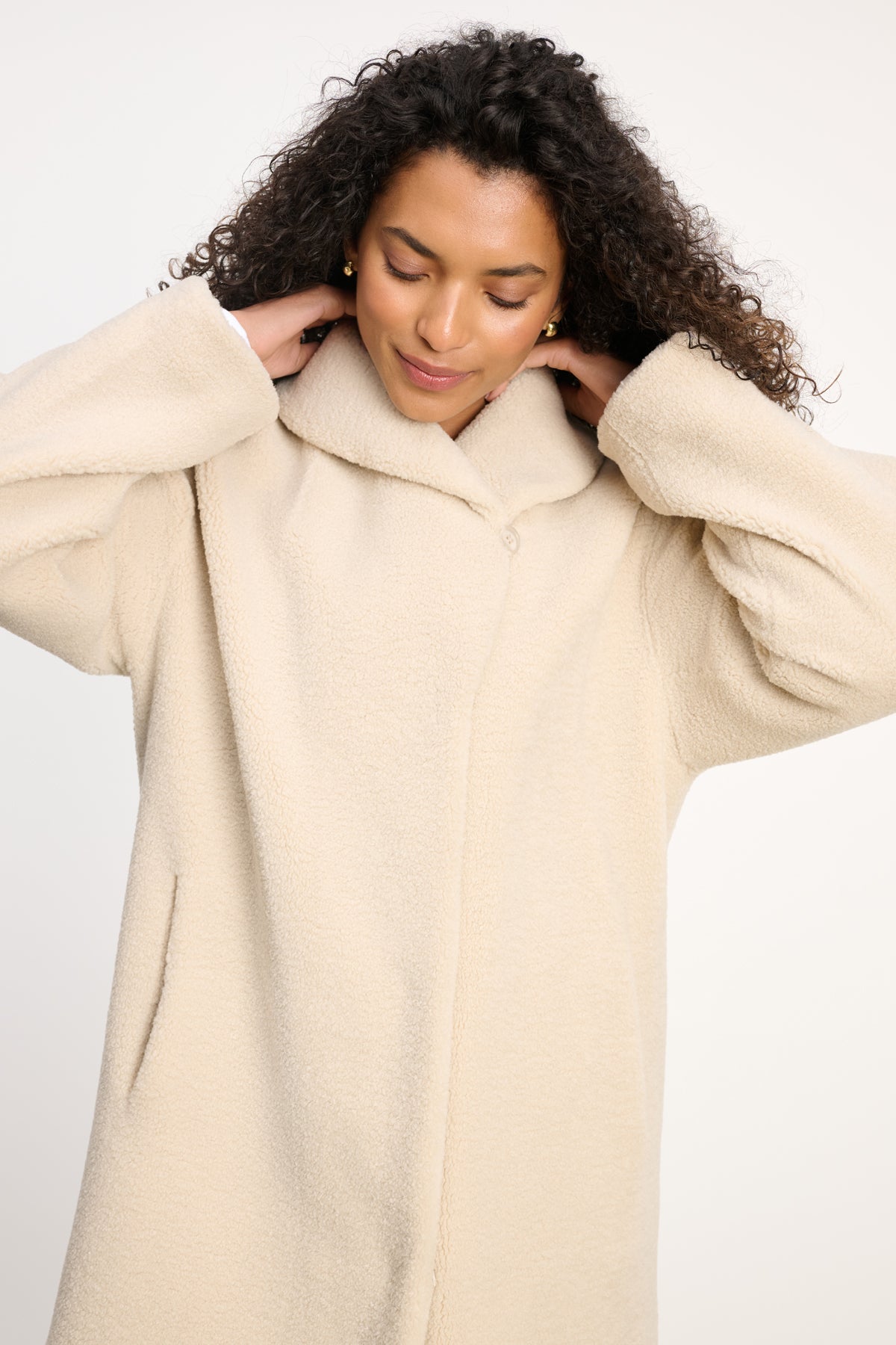 A person with curly hair wears the Velvet by Graham & Spencer ELEANOR SHERPA REVERSIBLE COAT in light beige, standing against a plain background and looking down with hands on the back of their neck.-39838347985089