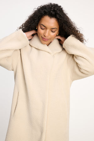 A person with curly hair wears the Velvet by Graham & Spencer ELEANOR SHERPA REVERSIBLE COAT in light beige, standing against a plain background and looking down with hands on the back of their neck.