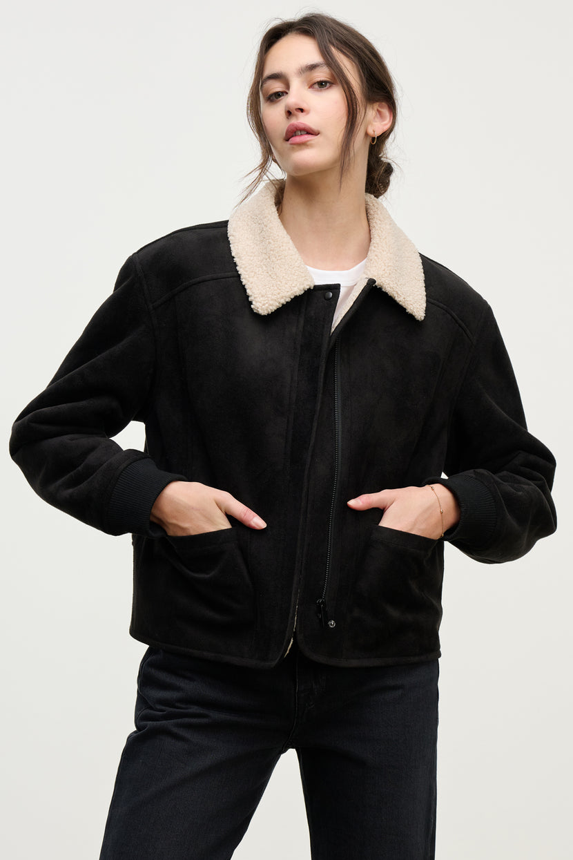 A person wears the JUNO SHERPA JACKET by Velvet by Graham & Spencer, featuring a black faux sherpa body with a beige collar, full zip front, paired with a white top and black pants, hands in pockets against a plain background for added warmth.