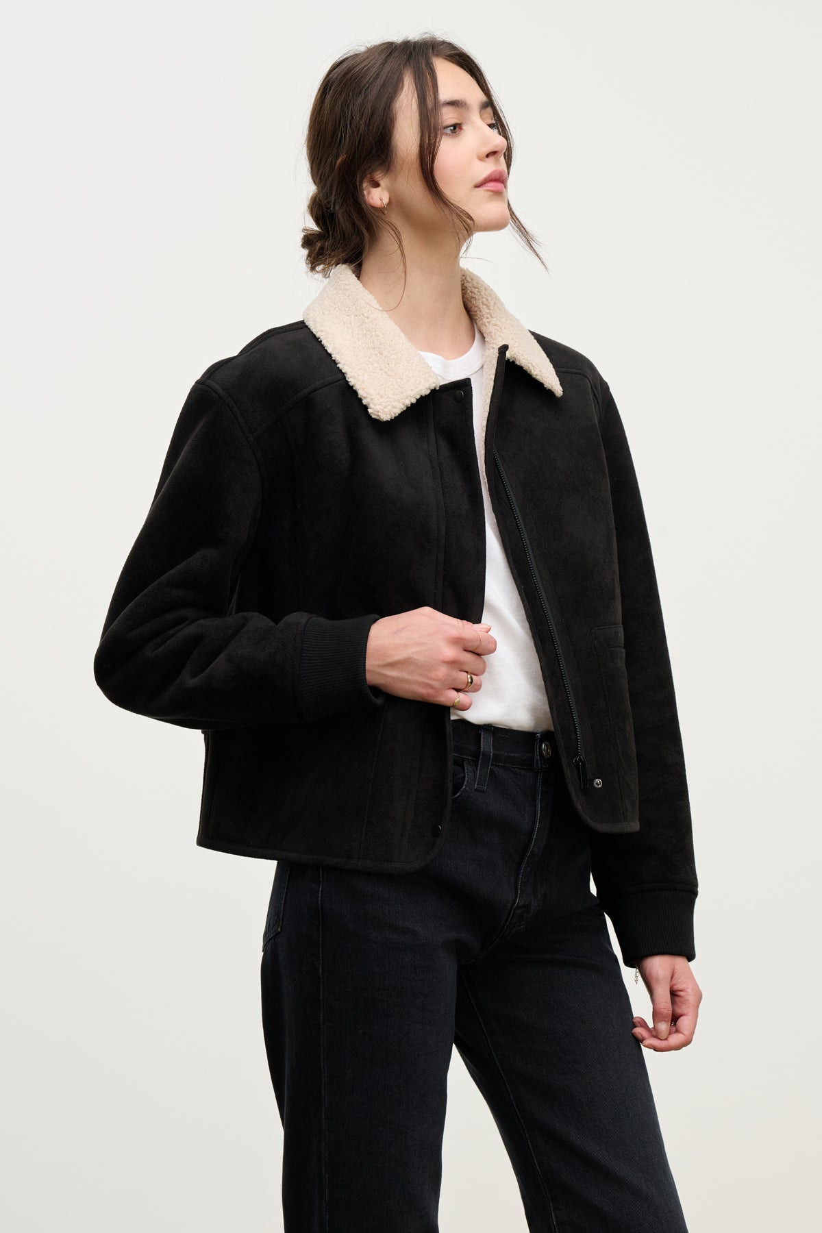 A person stands against a plain light background wearing the Velvet by Graham & Spencer JUNO SHERPA JACKET in black, styled with a white shirt and black jeans, highlighting its elevated warmth and style.-39838352801985