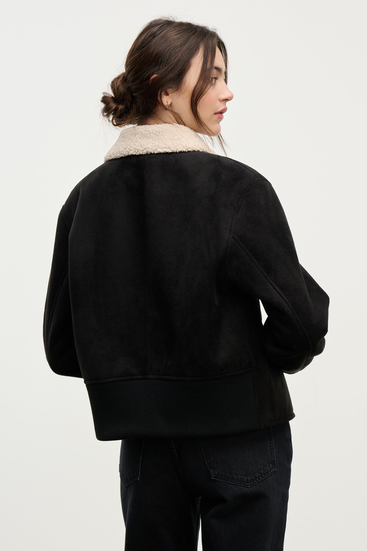 A woman with brown hair in a low bun is seen from behind wearing the JUNO SHERPA JACKET by Velvet by Graham & Spencer—a black faux sherpa jacket with a light-colored shearling collar, set against a plain background.-39838352769217