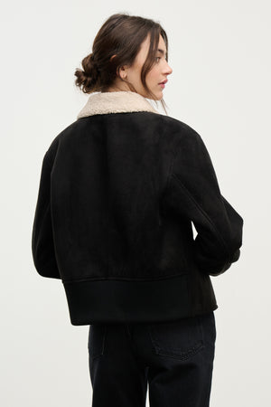 A woman with brown hair in a low bun is seen from behind wearing the JUNO SHERPA JACKET by Velvet by Graham & Spencer—a black faux sherpa jacket with a light-colored shearling collar, set against a plain background.