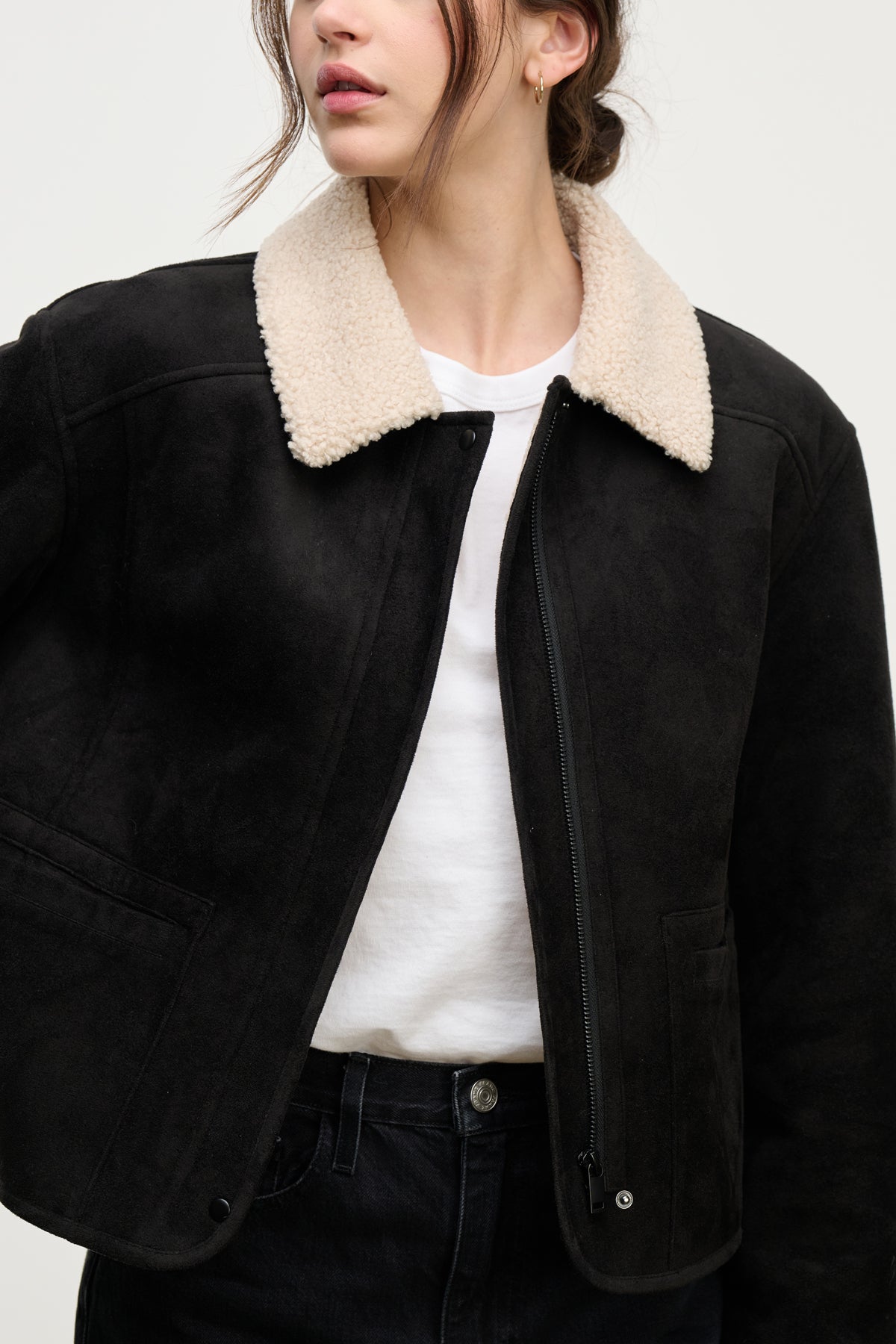 A woman wears the Velvet by Graham & Spencer JUNO SHERPA JACKET in black with a cream shearling collar, zipped over a white tee and black jeans. The full zip front offers style and cozy warmth against a plain background.-39838352736449