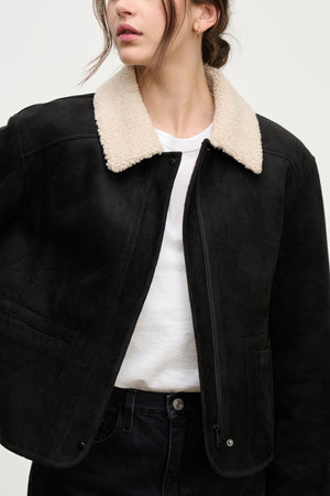 A woman wears the Velvet by Graham & Spencer JUNO SHERPA JACKET in black with a cream shearling collar, zipped over a white tee and black jeans. The full zip front offers style and cozy warmth against a plain background.