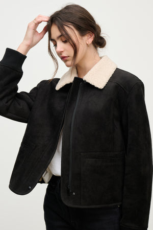 A person in the JUNO SHERPA JACKET by Velvet by Graham & Spencer, featuring a black faux sherpa body and light shearling collar over a white shirt, stands against a plain background, showcasing elevated warmth and effortless style.
