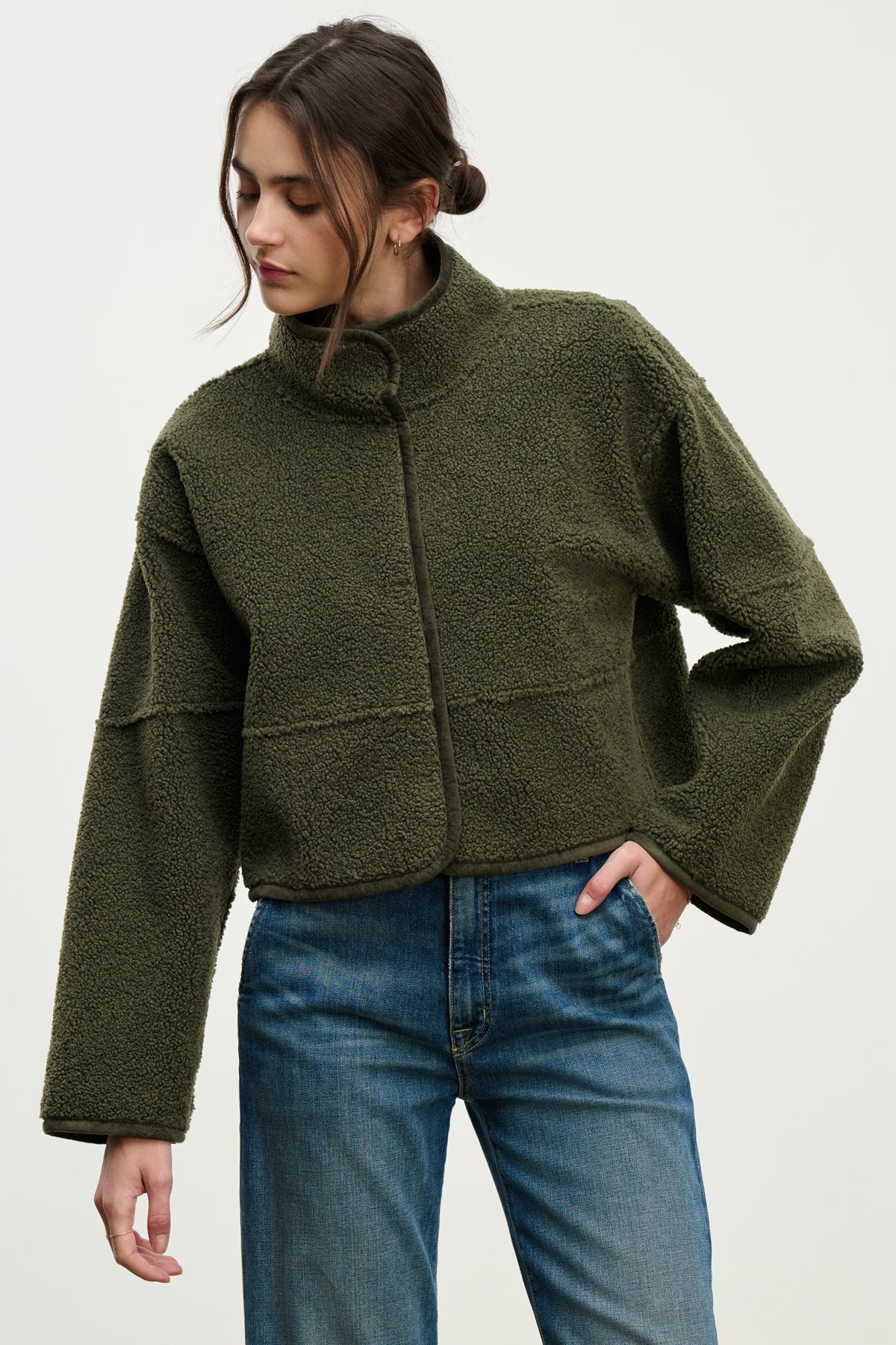 A person wearing the KELLY LUXE SHERPA REVERSIBLE JACKET by Velvet by Graham & Spencer in olive green, paired with blue jeans, stands against a plain light background, looking down to the side.-39800352112833