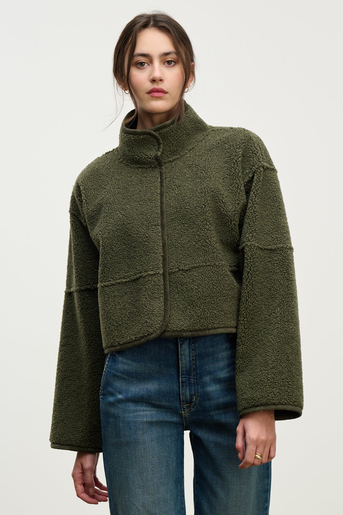   A woman wears the KELLY LUXE SHERPA REVERSIBLE JACKET by Velvet by Graham & Spencer—an olive green, cropped faux sherpa fleece with wide sleeves—paired with blue jeans, standing against a plain light background. 