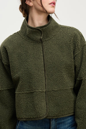 A person wearing the Velvet by Graham & Spencer KELLY LUXE SHERPA REVERSIBLE JACKET in olive green, featuring a high neck and textured faux sherpa fleece, paired with blue jeans and shown from shoulders to waist.