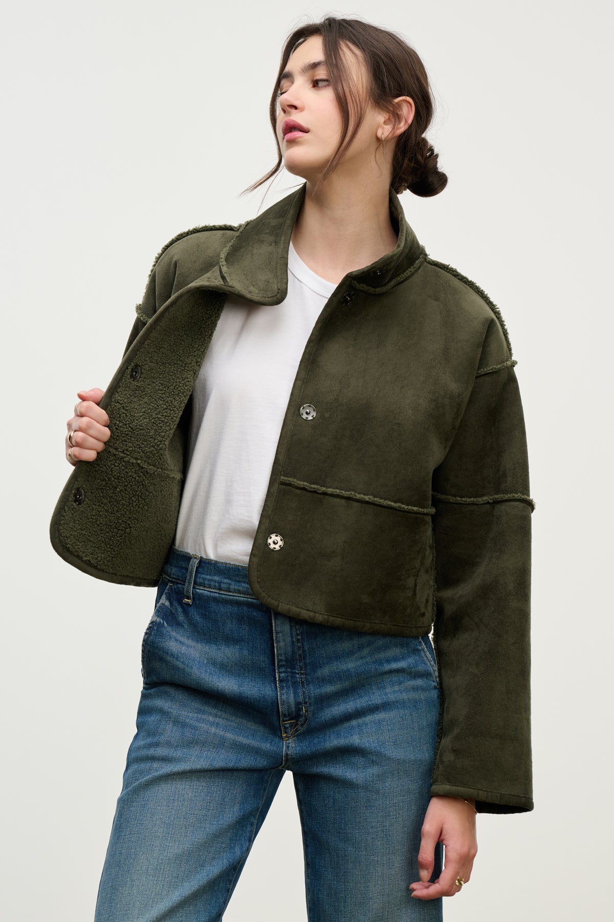 A person wears the Velvet by Graham & Spencer KELLY LUXE SHERPA REVERSIBLE JACKET in olive green, styled over a white t-shirt and blue jeans, standing against a plain light background.