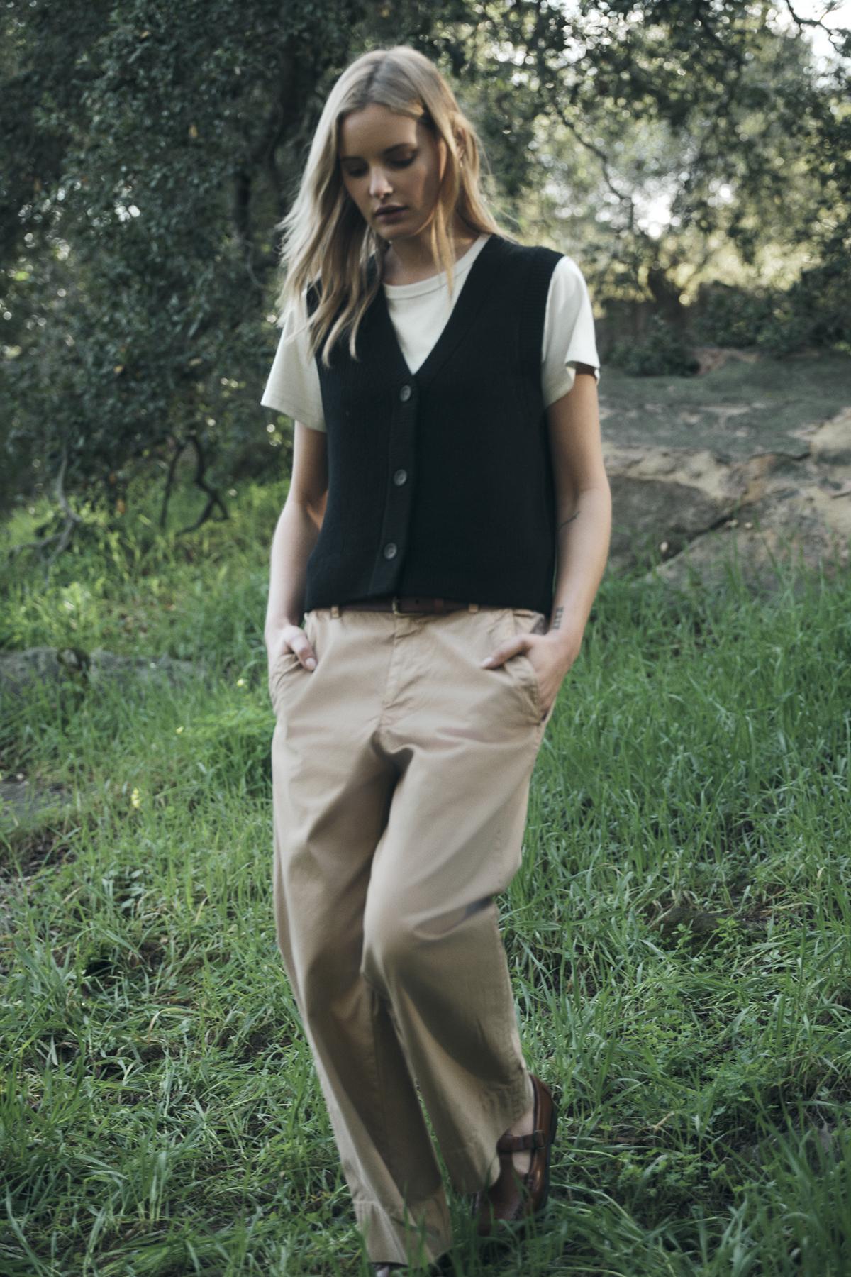   A person stands outdoors in casual attire, wearing a white V-neck t-shirt, the LU SWEATER VEST by Velvet by Graham & Spencer in black wool blend, and beige pants, with hands in pockets. Green grass and trees are in the background. 