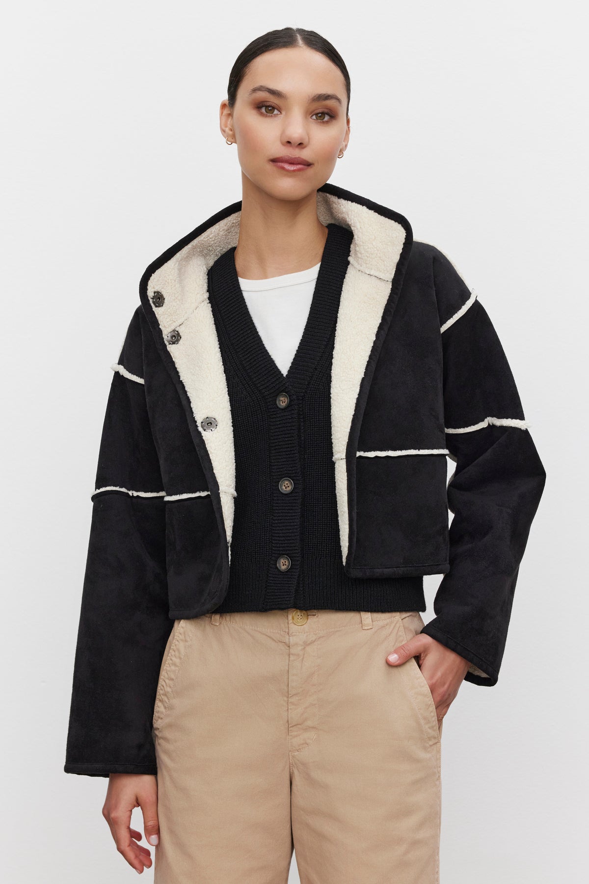   A person stands against a white background, wearing the black KELLY LUXE SHERPA REVERSIBLE JACKET from Velvet by Graham & Spencer over a black sweater and beige pants. 