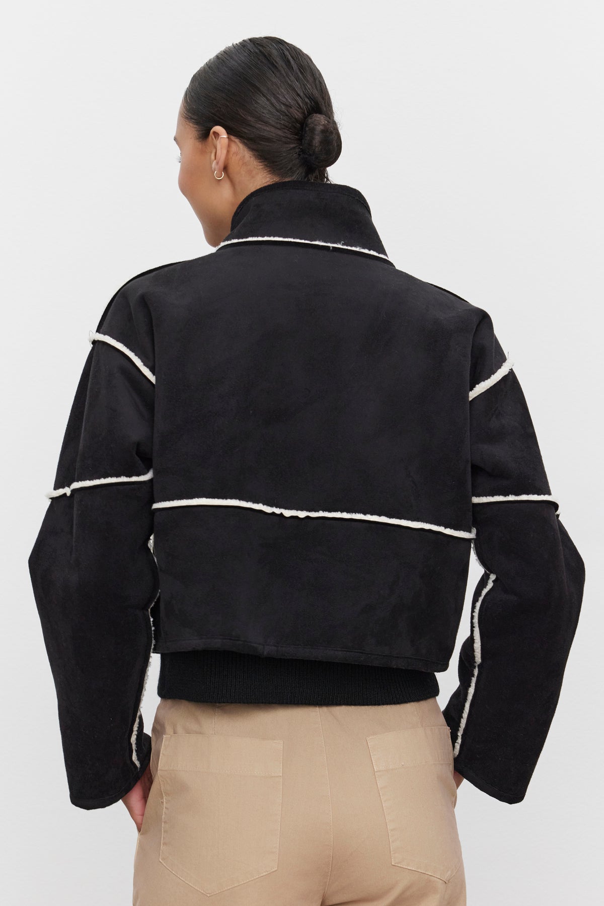   A person is wearing the KELLY LUXE SHERPA REVERSIBLE JACKET by Velvet by Graham & Spencer in black, along with tan pants, facing away from the camera to highlight the jacket's white faux sherpa trim. 